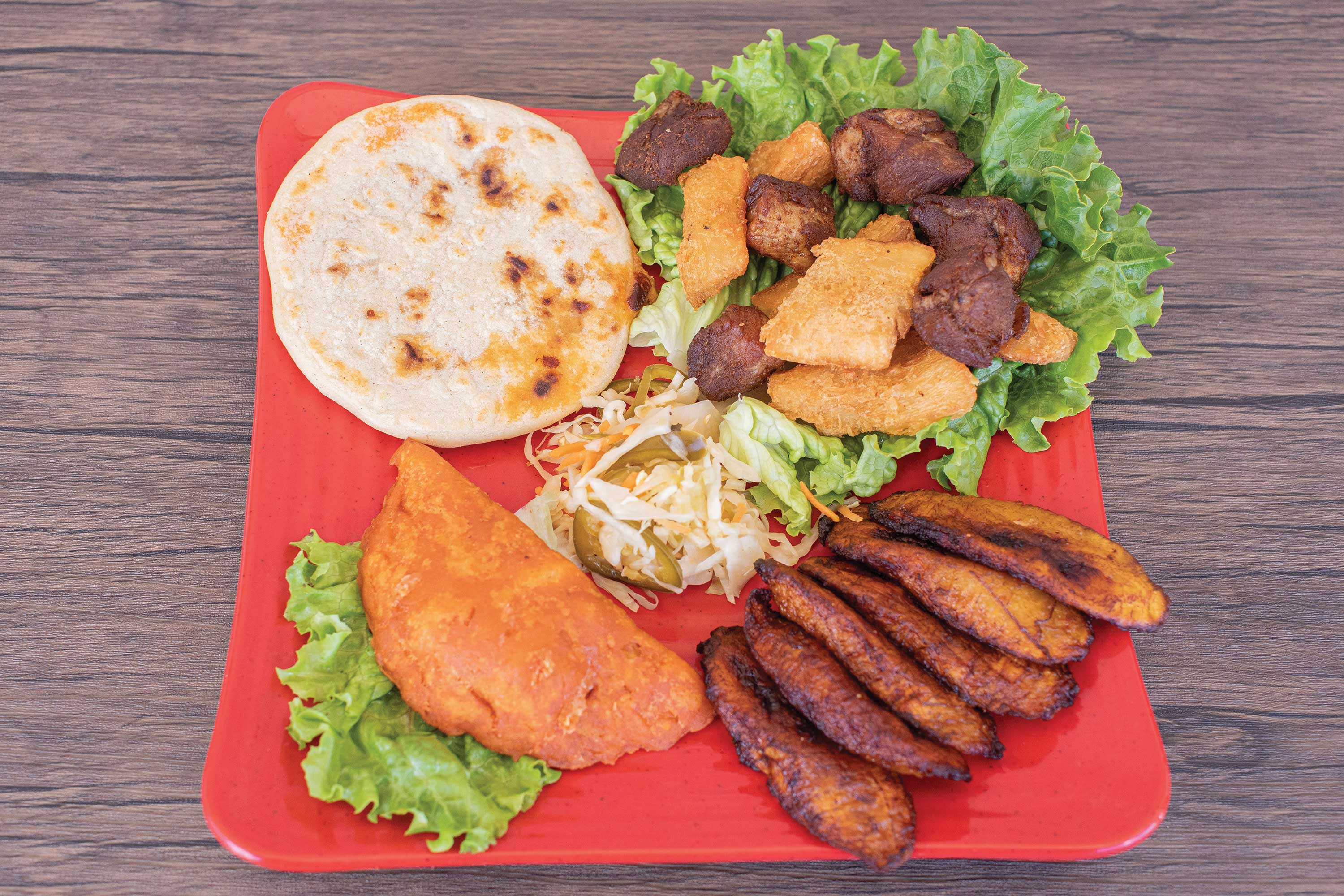 Image: A grilled pupusa, fanned out fried plantains, a pastelito turnover, and a mixture of meat and fried yucca served on a bed of lettuce on a red plate. A small mound of pickled cabbage is in the center of the plate.