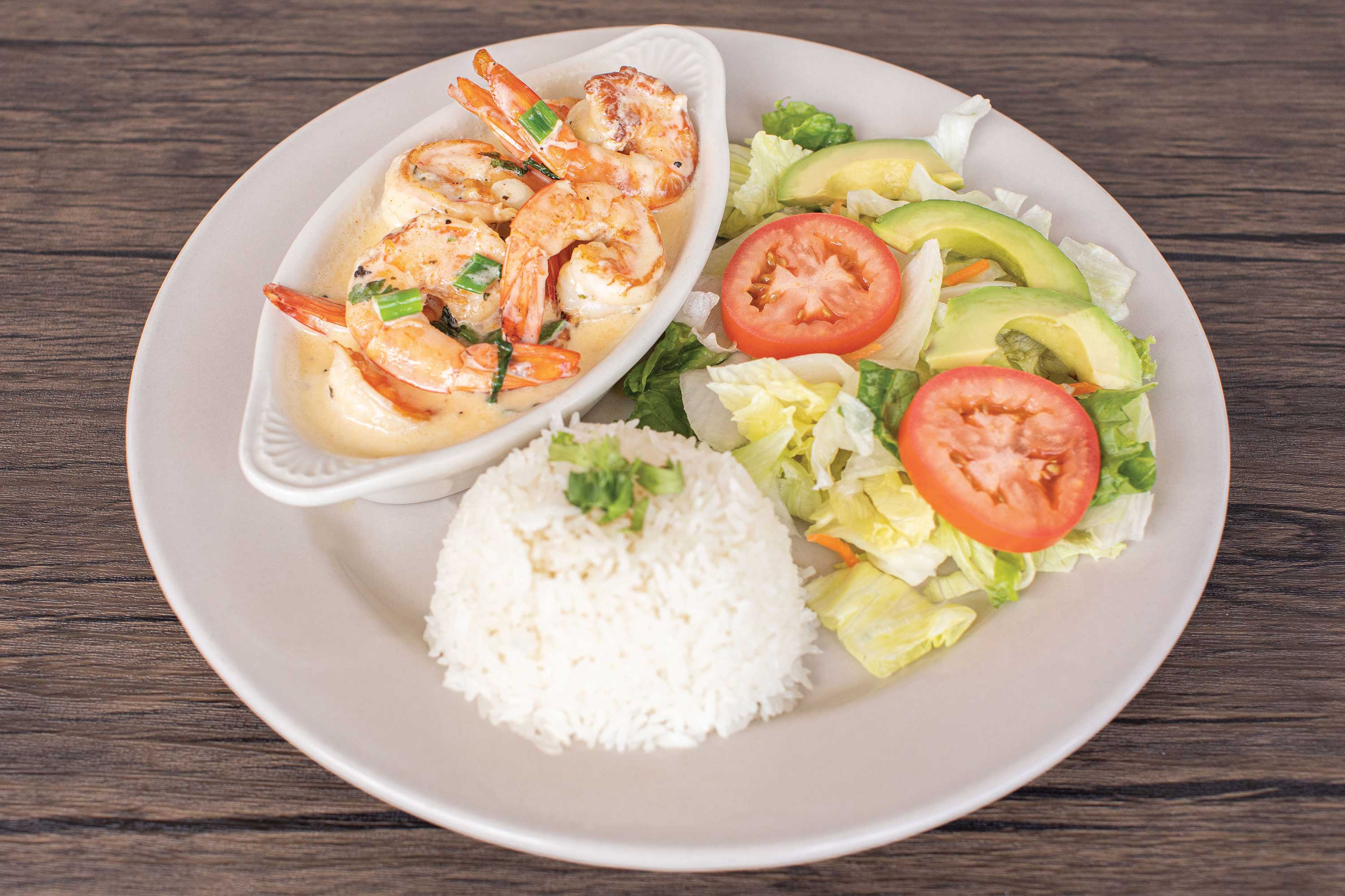 Image: Grilled shrimp in a creamy sauce in a white dish. Served alongside a lettuce and tomato salad with a mount of white rice.