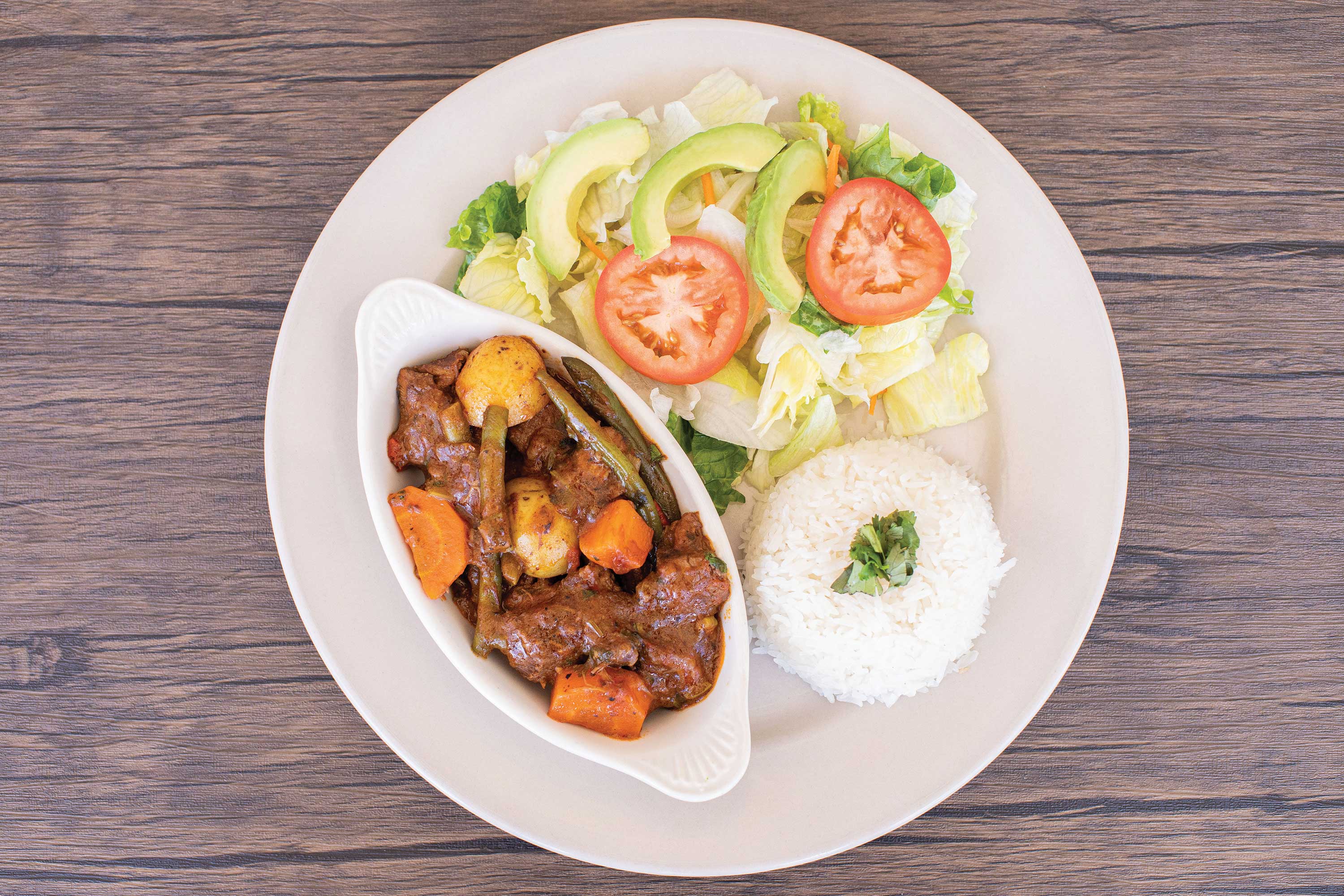 Image: Stewed beef with potato, greeen beans and carrots served in a white dish next to a lettuce and tomato salad and a mound of white rice.