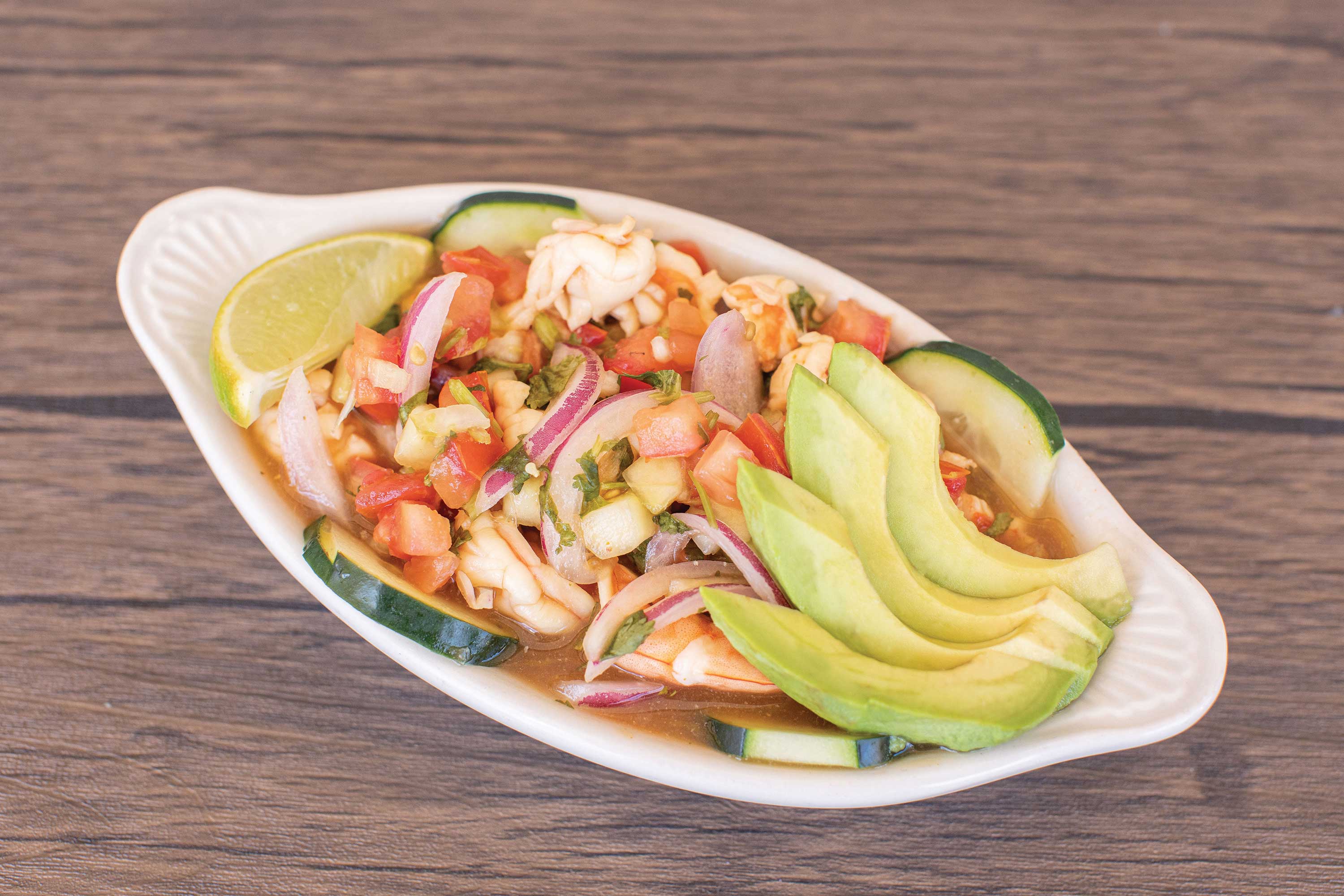 Image: Shrimp mixed with diced tomatoes, onions, celery and jalapenos in a white serving dish. Topped with chopped cilantro, sliced cucumber, slices of avocado and a lime wedge.