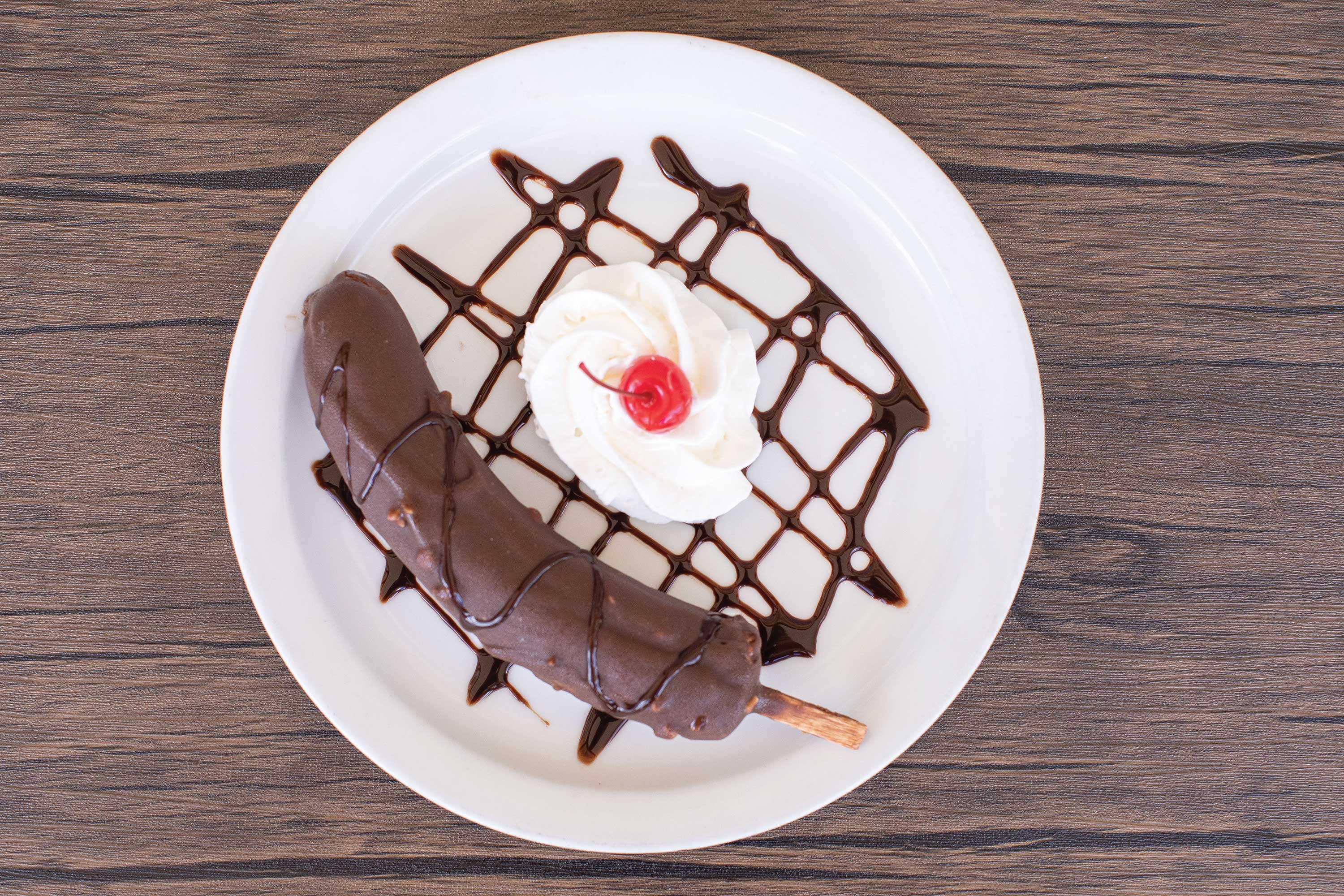 Image: A chocolate covered banana on a stick is set on a white plate alongside a mound of whipped cream topped with a cherry. Chocolate sauce is drizzled over the plate in a decorative pattern.