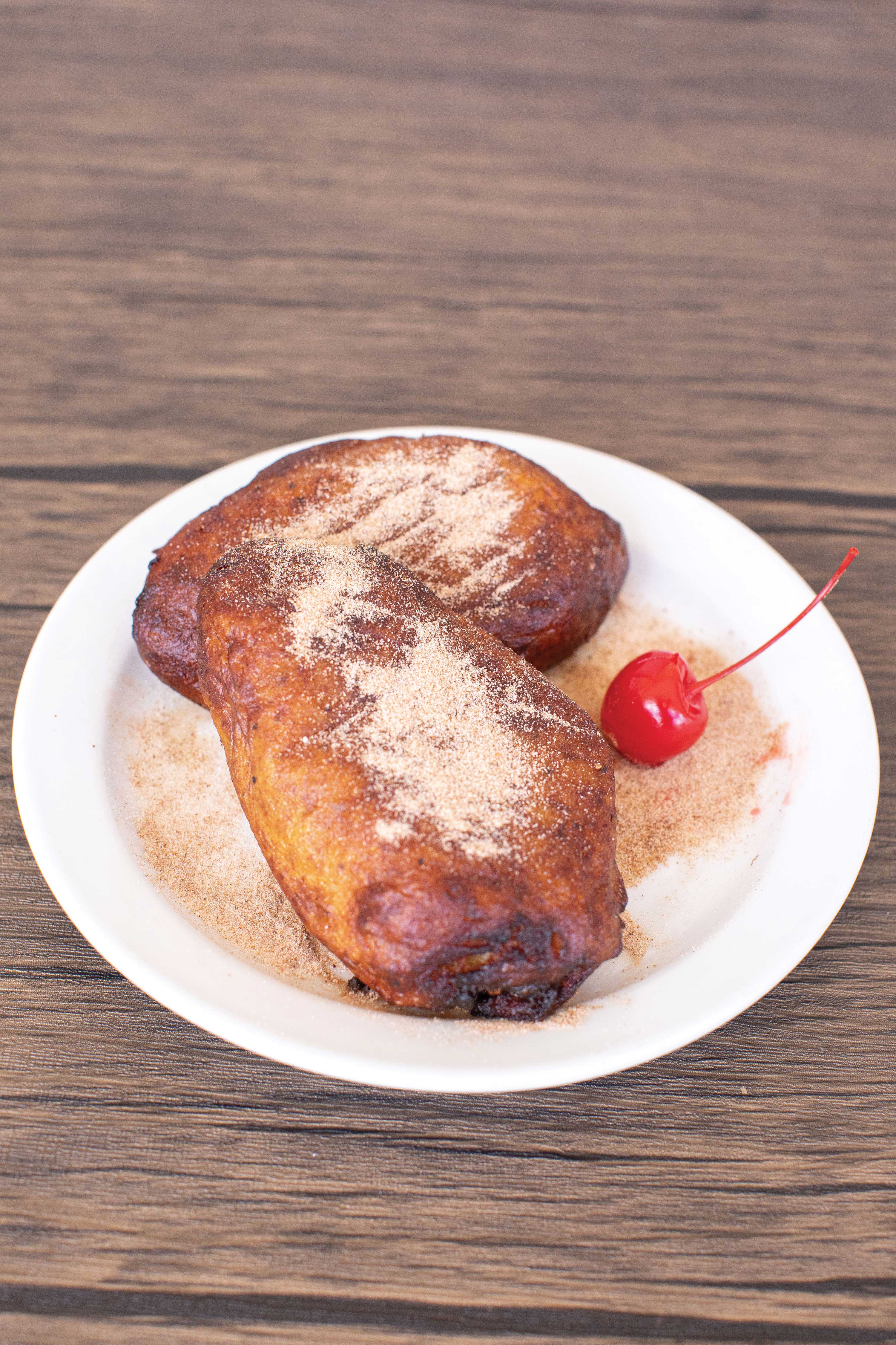 Image: Two brown turnovers are set on a white plate. They are sprinkled with cinnamon sugar and topped with a cherry.