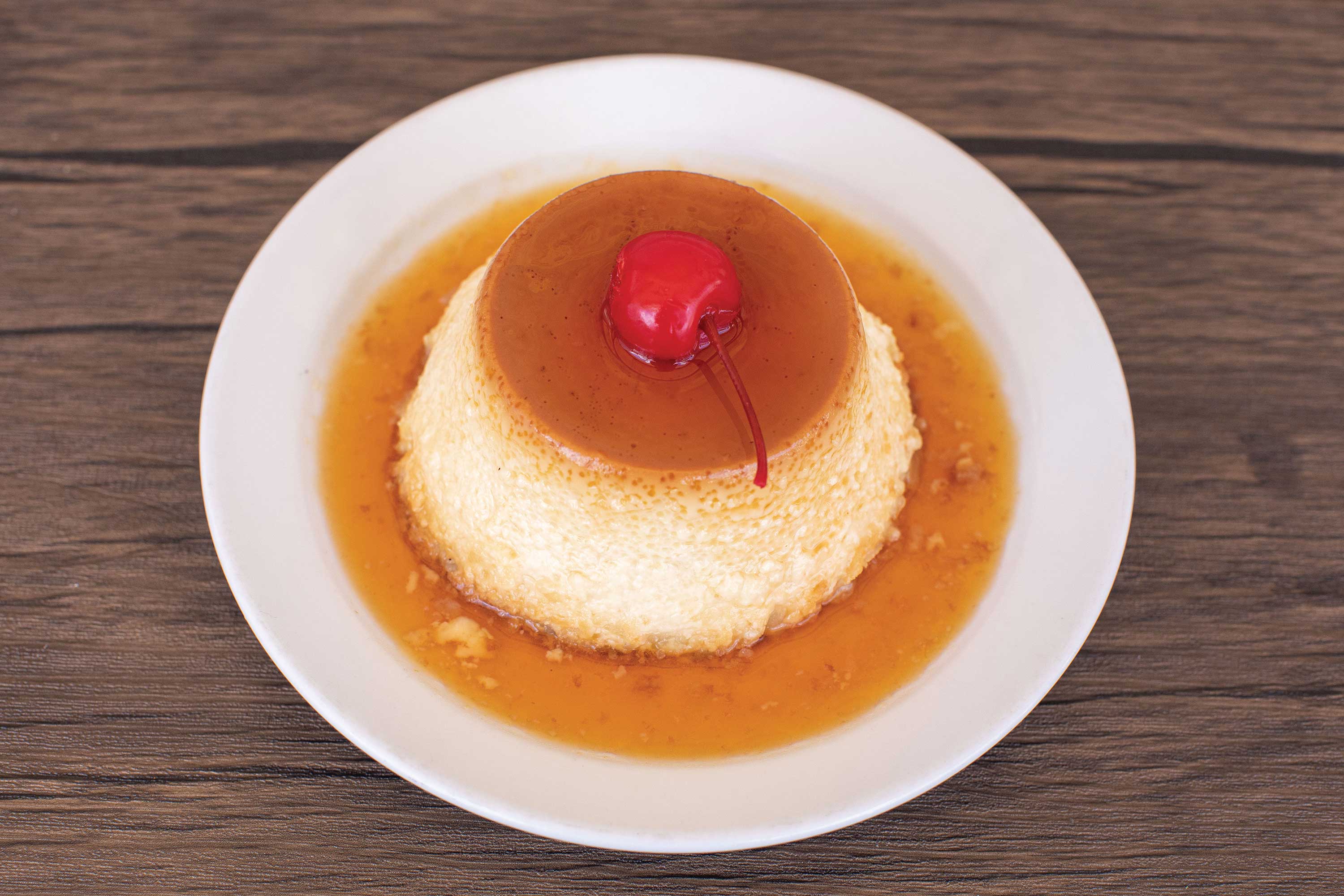 Image: A flan custard covered in a thin caramel sauce is served on a white plate. A cherry is set on top.