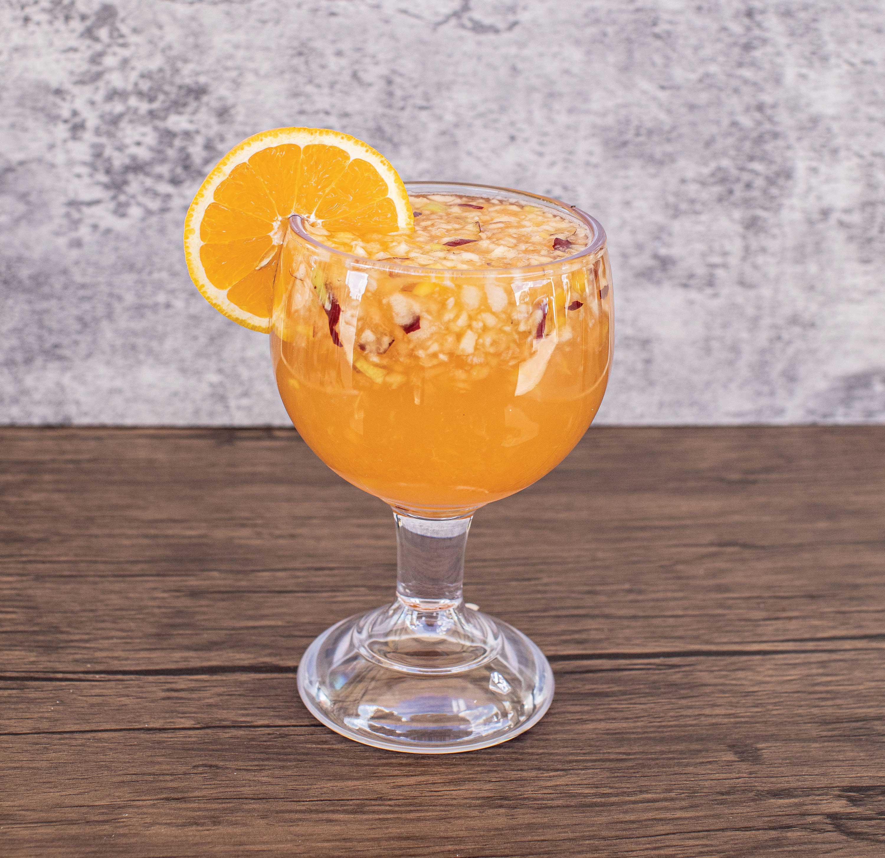 Image: A large glass is filled with an orange liquid mixed with bits of fruit. An orange slice is set on the glass rim.