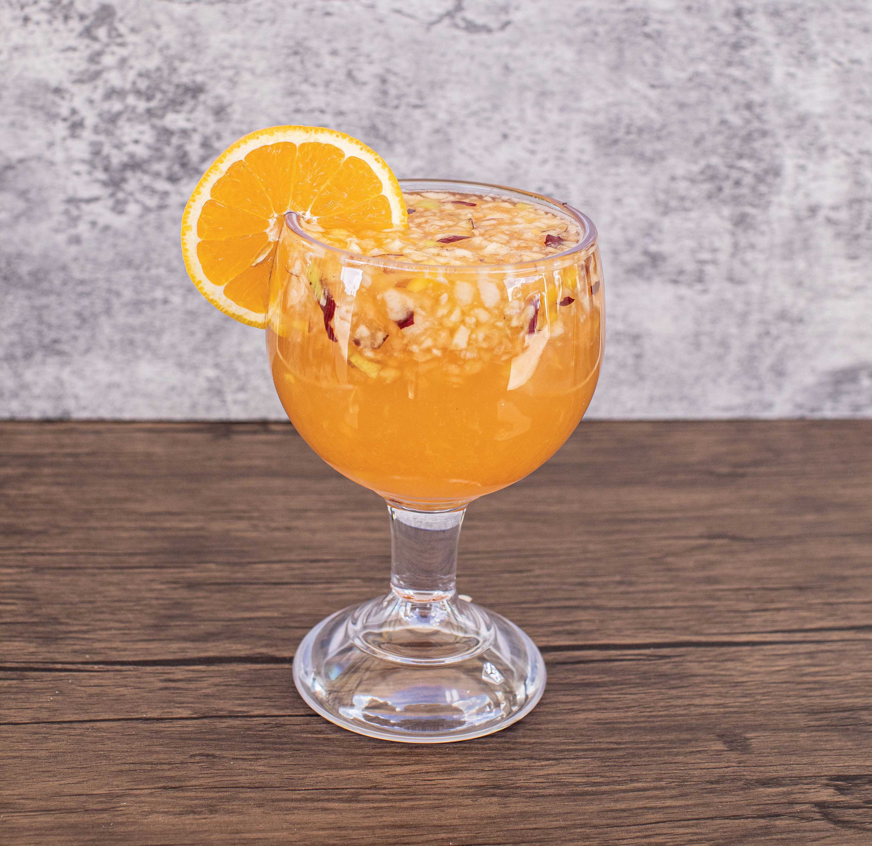 Image: A large glass is filled with an orange liquid mixed with bits of fruit. An orange slice is set on the glass rim.
