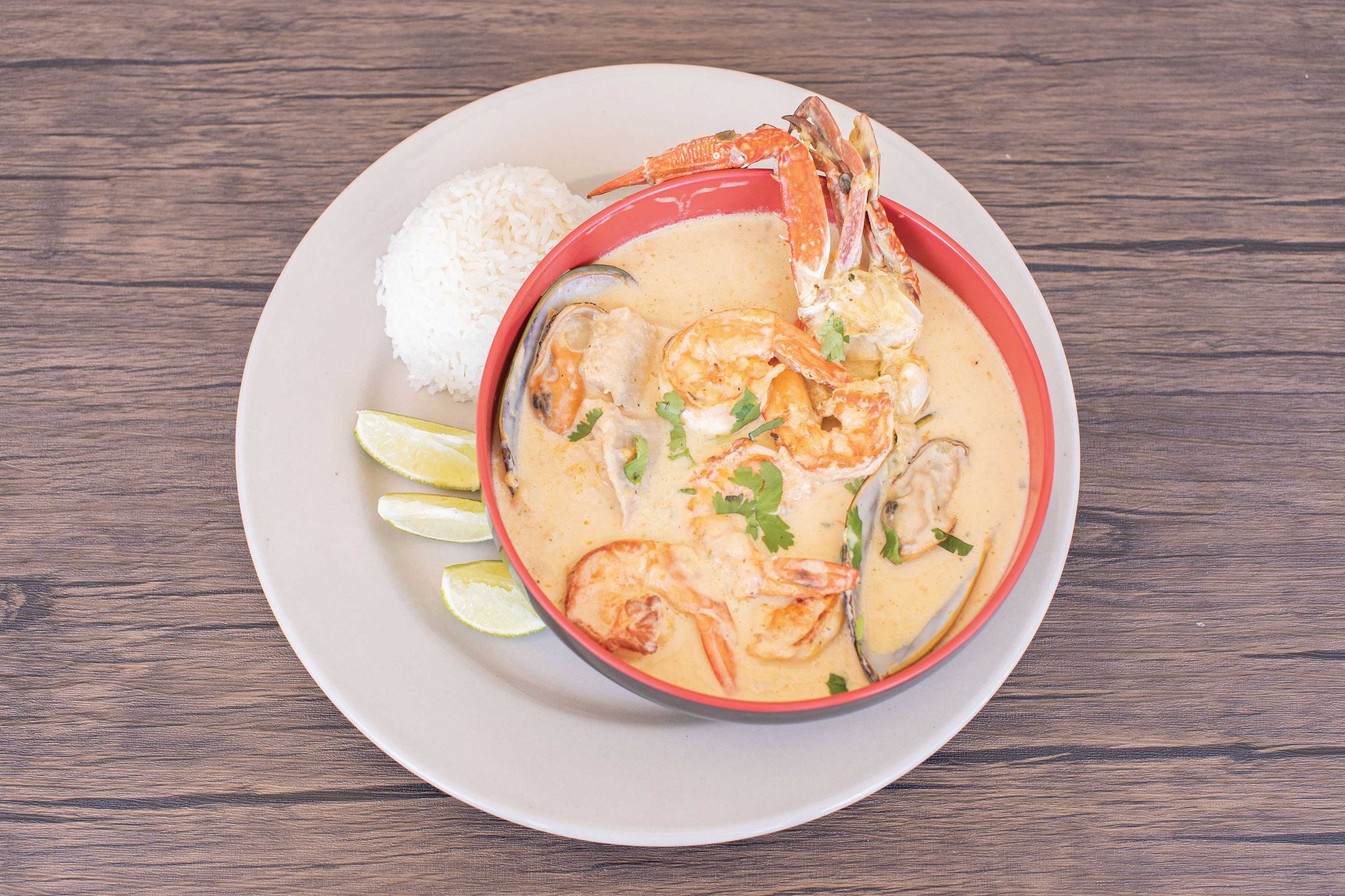 Image: Red bowl filled with a creamy soup. Shrimp, mussels in shell, and crab are arranged on top with a sprinkling of cilantro. Served alongside lime wedges and a mound of white rice.