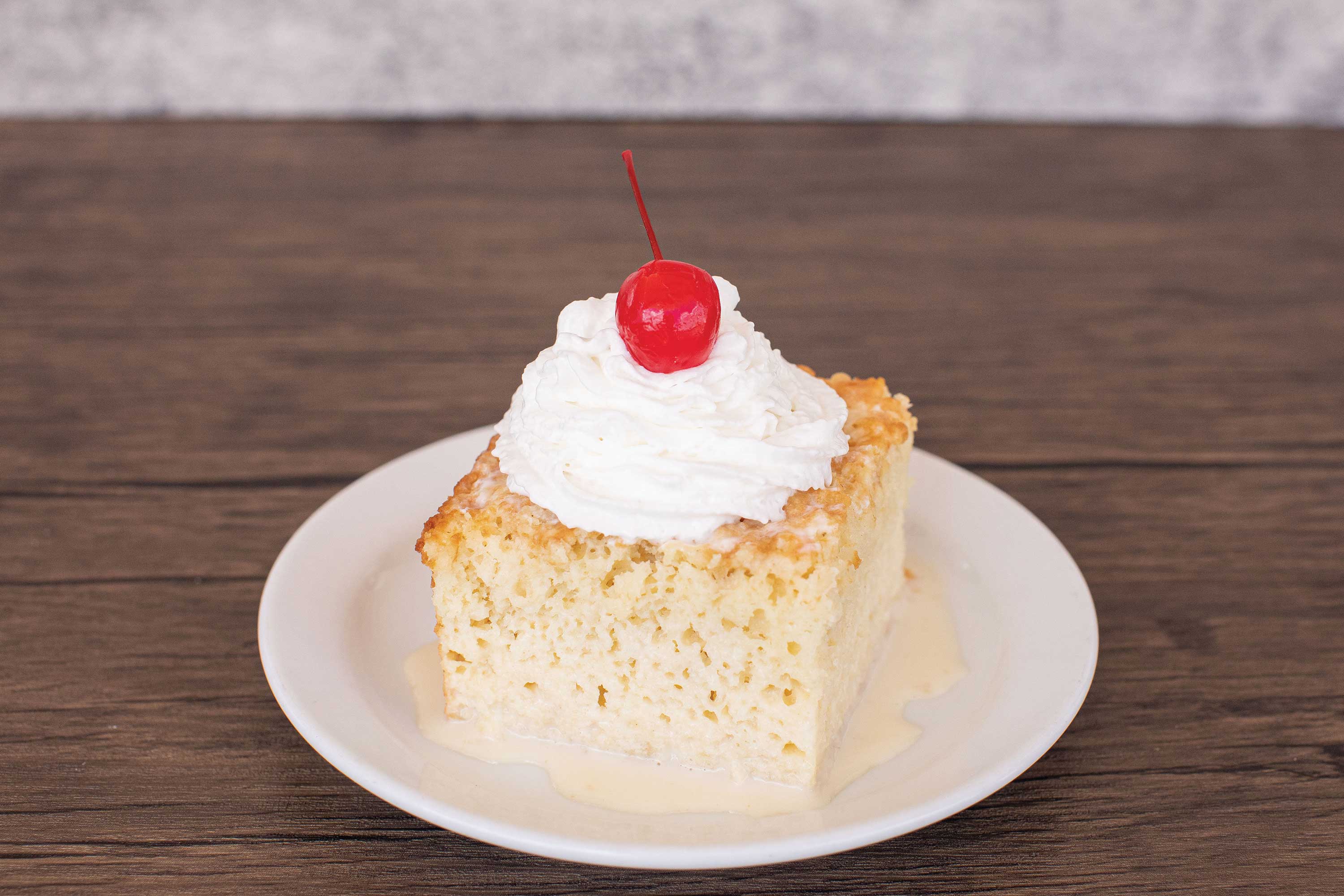 Image: A square slice of cake is set on a white plate. The cake is moist and sits in a small pool of white sauce. Whipped cream and a cherry sits on top of the cake.